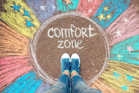 Pessoa a caminhar numa estrada entre montanhas com a frase 'Comfort Zone' pintada no asfalto, simbolizando a jornada para sair da zona de conforto.