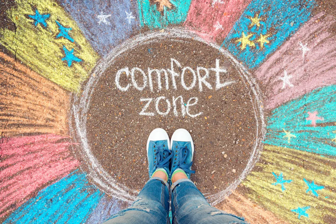 Pessoa a caminhar numa estrada entre montanhas com a frase 'Comfort Zone' pintada no asfalto, simbolizando a jornada para sair da zona de conforto.