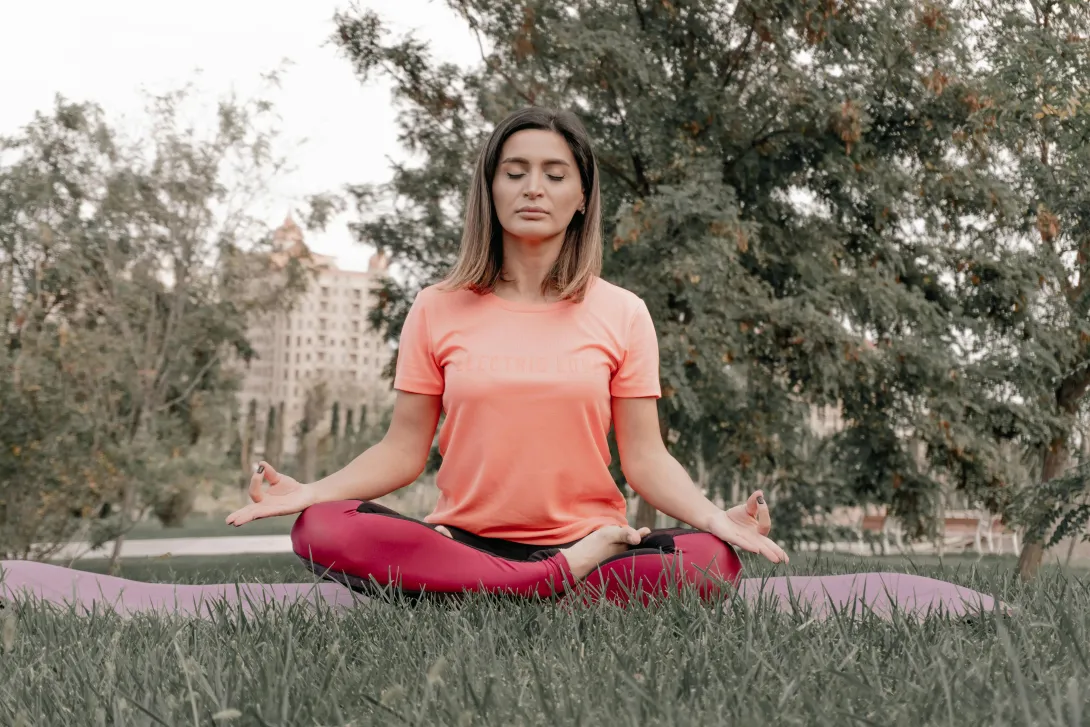 Mulher sentada em posição de lótus no parque, a meditar com serenidade, simbolizando equilíbrio e transformação pessoal.